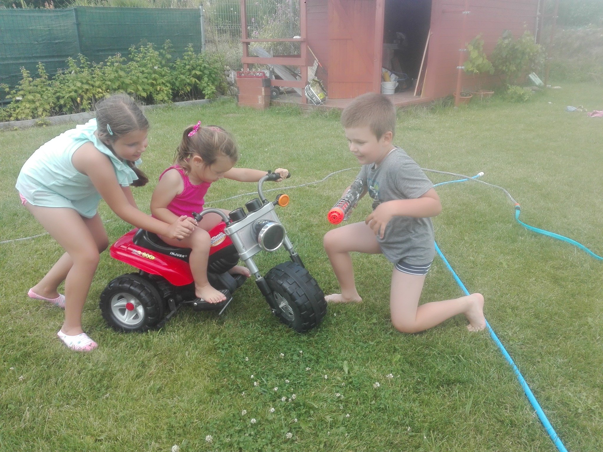 Dwoje dzieci pomaga dziewczynce z kokardą na włosach jeździć na czerwonym, plastikowym quadzie na trawniku przed drewnianą szopą.