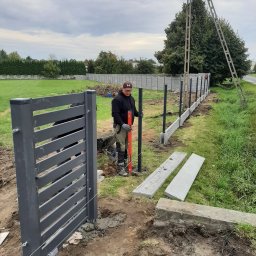 Hermes ogrodzenia - Montaż nowoczesnej, szarej bramy i słupków ogrodzeniowych na posesji; w tle widoczny fragment betonowego ogrodzenia i pracownik z poziomicą.