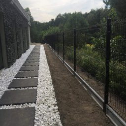 Hermes ogrodzenia - Czarny metalowy płot panelowy z betonową podmurówką, oddzielający posesję od zalesionego terenu, obok ścieżka z betonowych płyt otoczonych białymi kamykami.