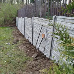 Szary, prefabrykowany mur z cegieł, w tle brązowe ogrodzenie z metalowych prętów, dwie łopaty oparte o przęsło, ziemia po montażu, trawa i krzewy.