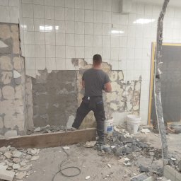 Robert Urbański Hermes - Demontaż płytek ściennych w łazience podczas remontu. Widoczny fragment ściany z resztkami kleju, gruz na podłodze i pracownik w trakcie pracy.