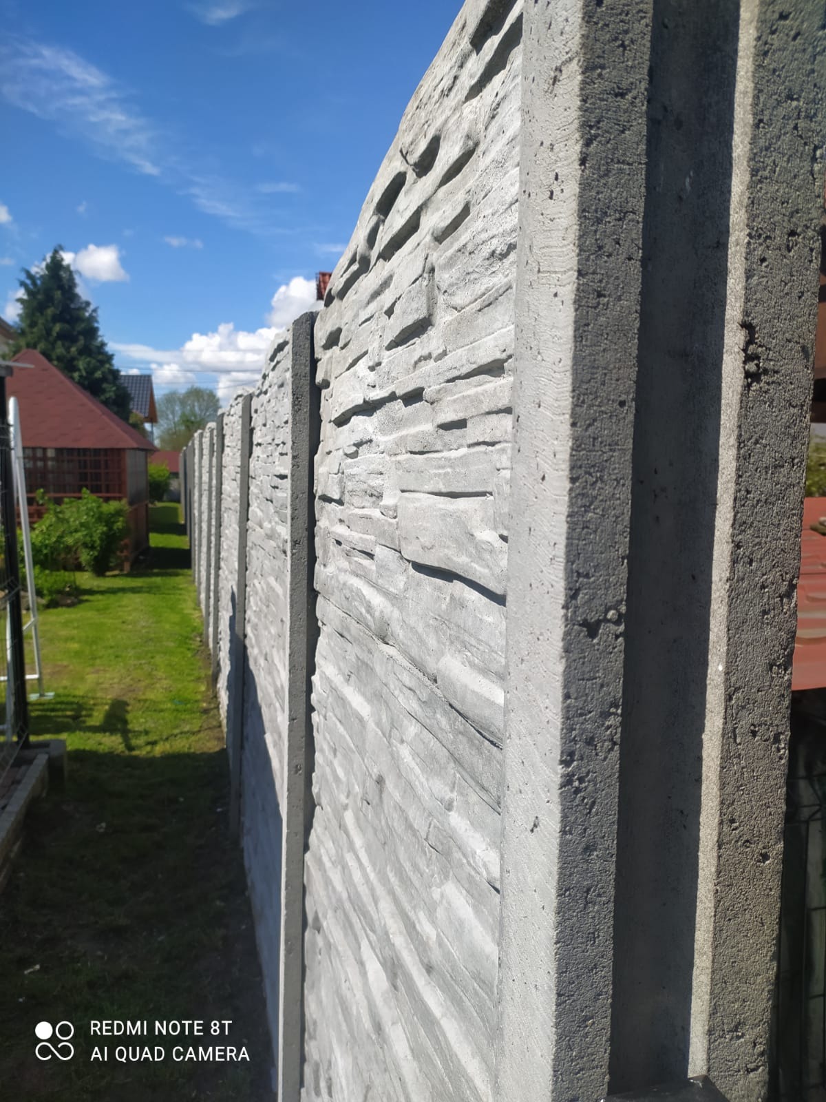 Długi, szary mur z prefabrykowanych elementów betonowych imitujących kamień, tworzący ogrodzenie na tle zielonego trawnika i zabudowań mieszkalnych pod błękitnym niebem.