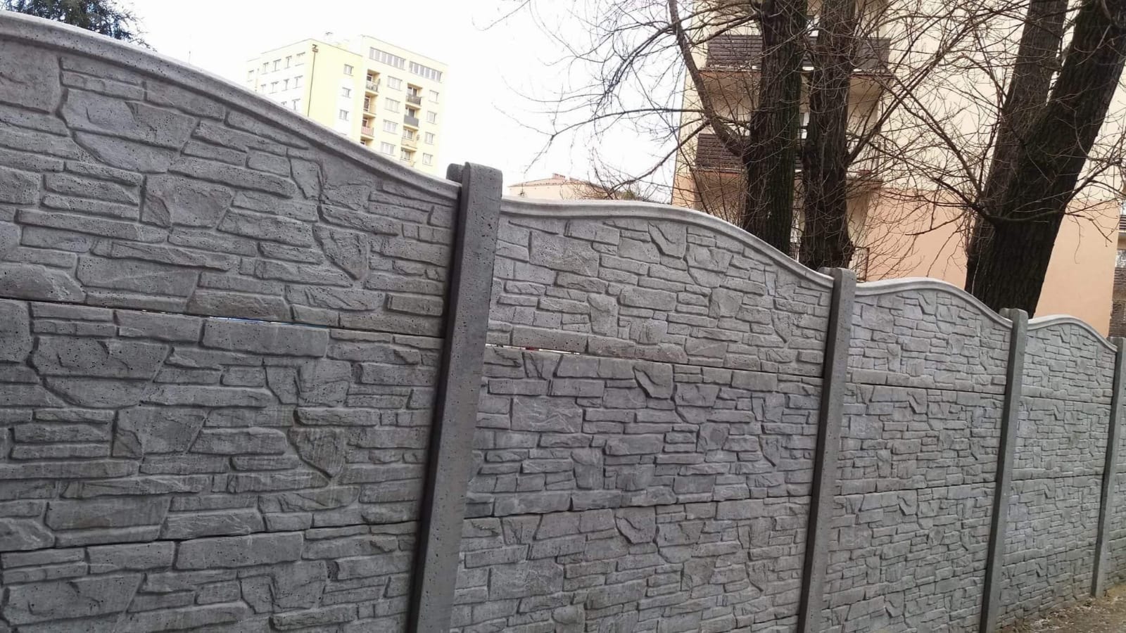 Szare, prefabrykowane ogrodzenie betonowe imitujące kamień, z widocznymi łączeniami paneli i słupkami, na tle drzew i budynku.