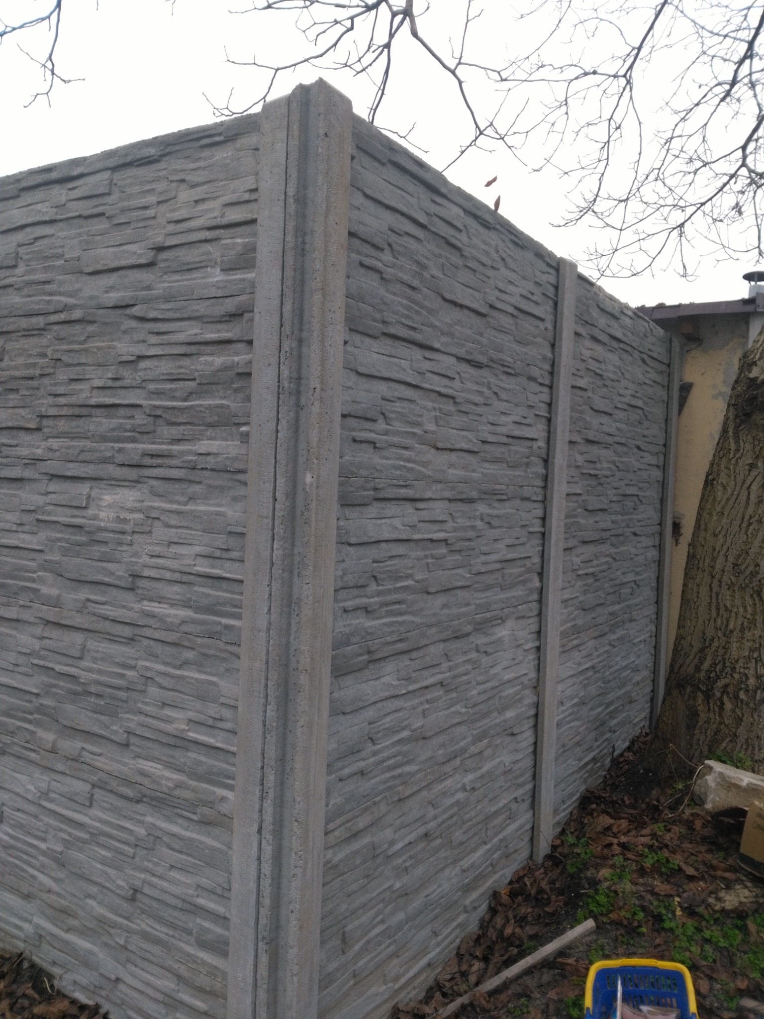 Szary, prefabrykowany płot z betonu imitującego kamień, z widocznymi słupkami, ustawiony na ziemi pokrytej liśćmi, z fragmentem drzewa i budynku w tle.