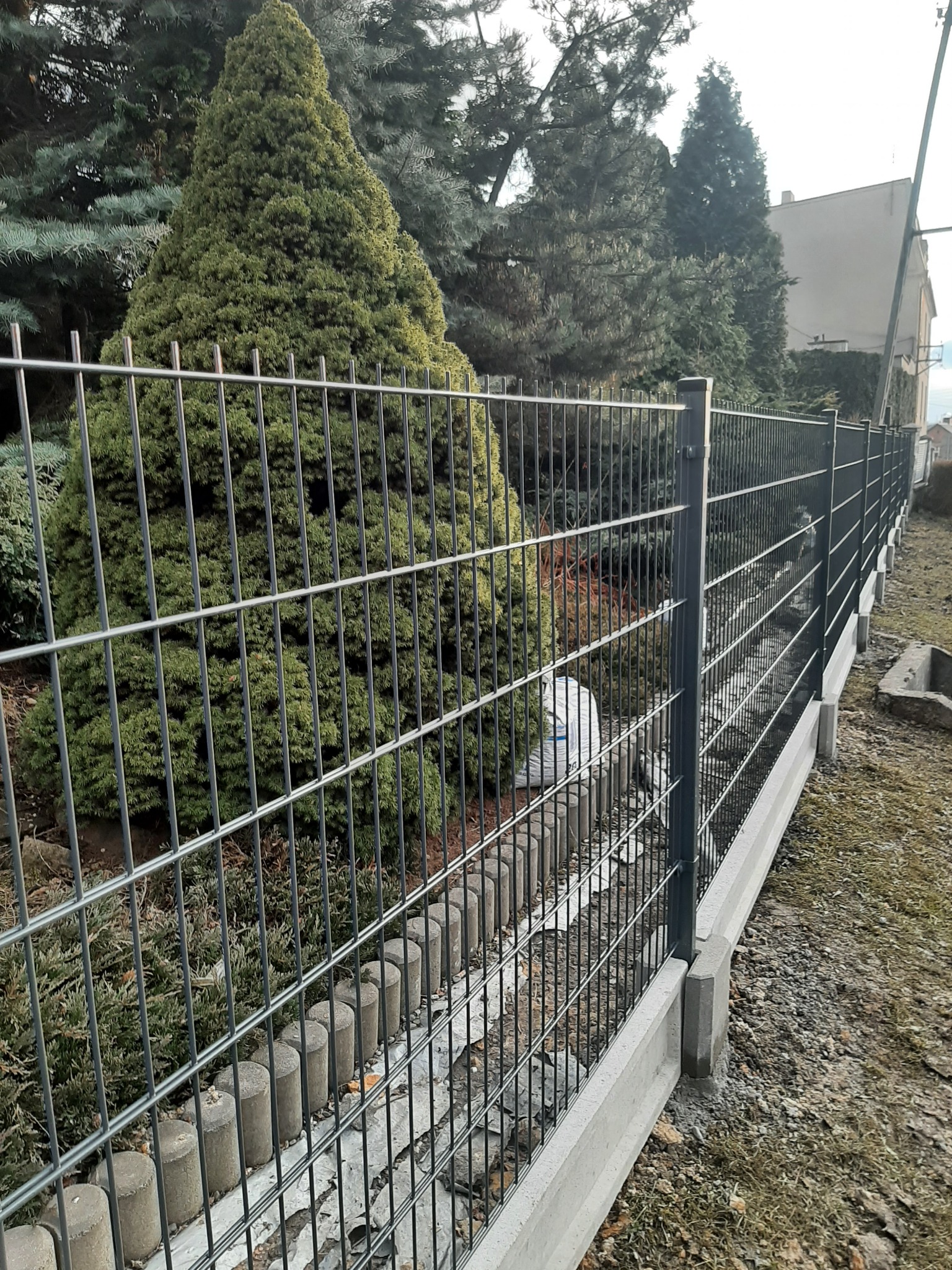 Długie, metalowe ogrodzenie panelowe z podmurówką betonową i elementami dekoracyjnymi z betonu, widok z zewnątrz posesji, w tle zielone drzewa iglaste.