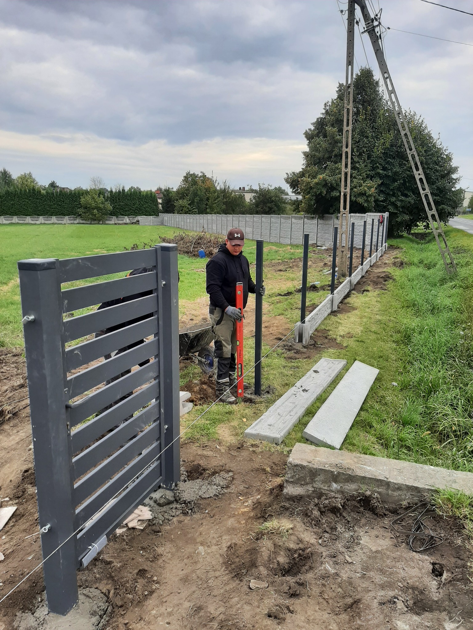 Montaż nowoczesnej, szarej bramy i słupków ogrodzeniowych na posesji; w tle widoczny fragment betonowego ogrodzenia i pracownik z poziomicą.