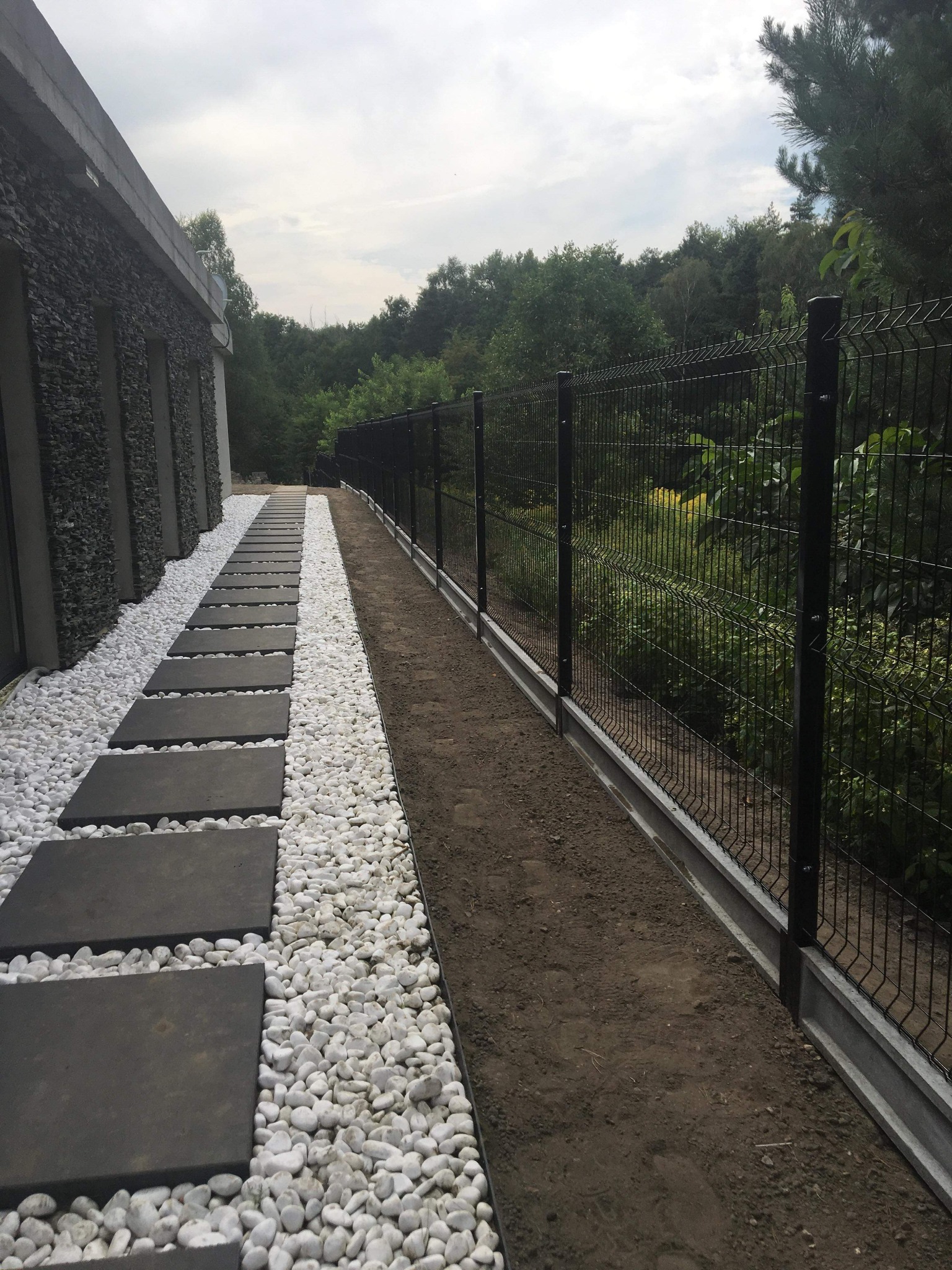 Czarny metalowy płot panelowy z betonową podmurówką, oddzielający posesję od zalesionego terenu, obok ścieżka z betonowych płyt otoczonych białymi kamykami.