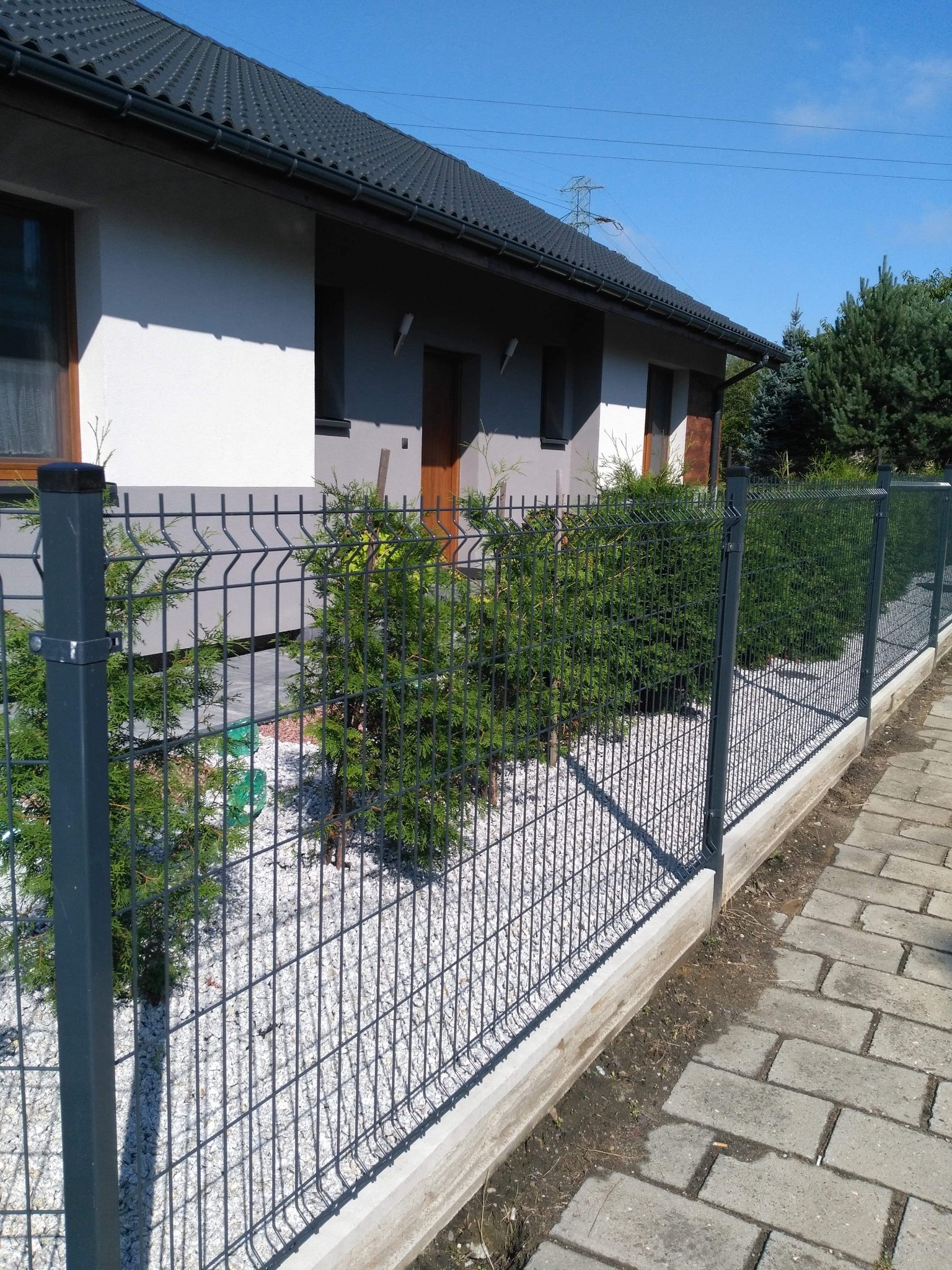 Nowoczesne ogrodzenie panelowe z metalu, z podmurówką betonową i białym grysem, oddzielające posesję z domem jednorodzinnym, z widoczną kostką brukową chodnika.