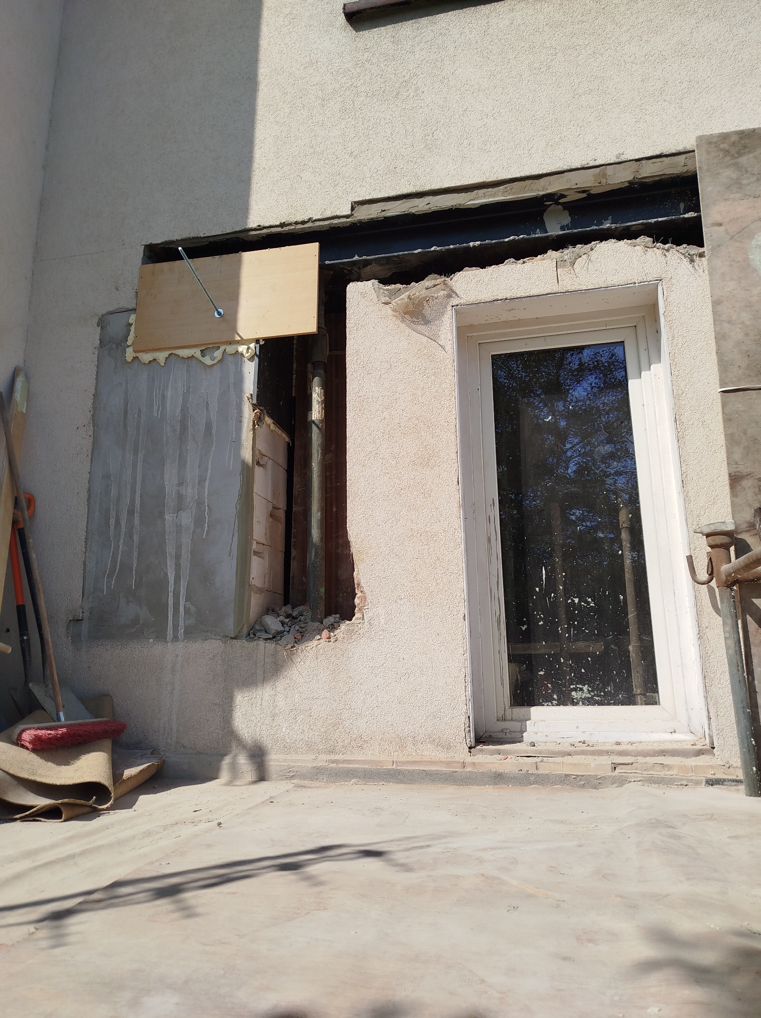 Otwór okienny w trakcie prac murarskich, częściowo zamurowany z widoczną drewnianą płytą mocującą, narzędzia murarskie obok.