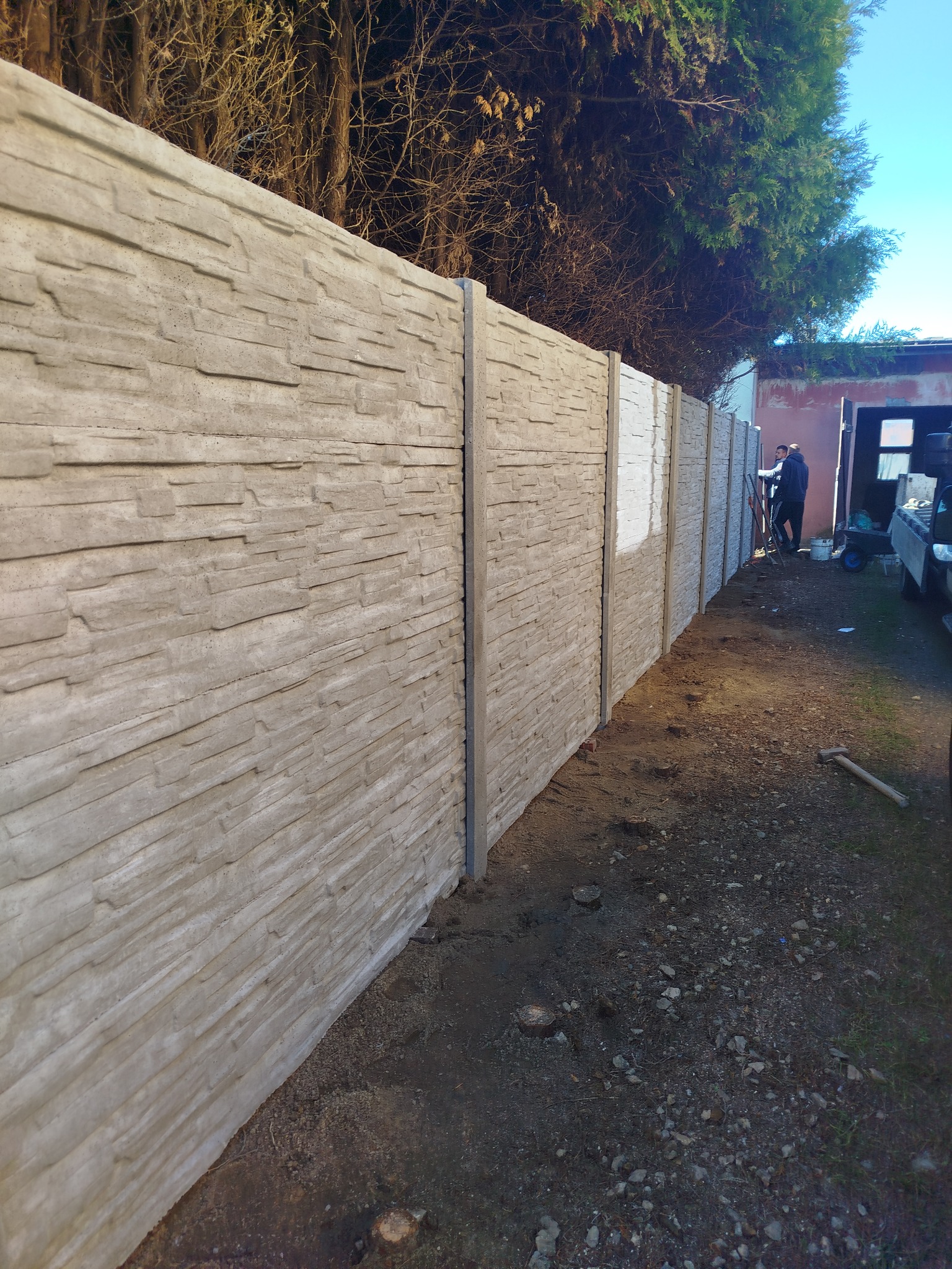Długi, nowo postawiony, szary płot z prefabrykatów betonowych imitujących kamień, widok z boku na nierówny teren, w tle sylwetki dwóch osób podczas pracy.