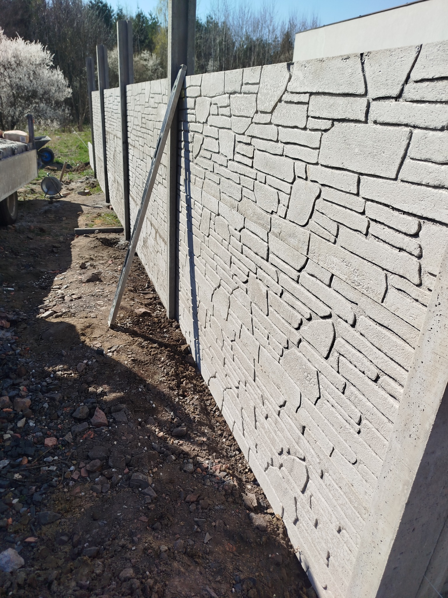 Nowo postawiony szary płot z betonu imitującego kamień, z widocznymi słupkami i łatą murarską opartą o konstrukcję, teren wokół w trakcie prac ziemnych.