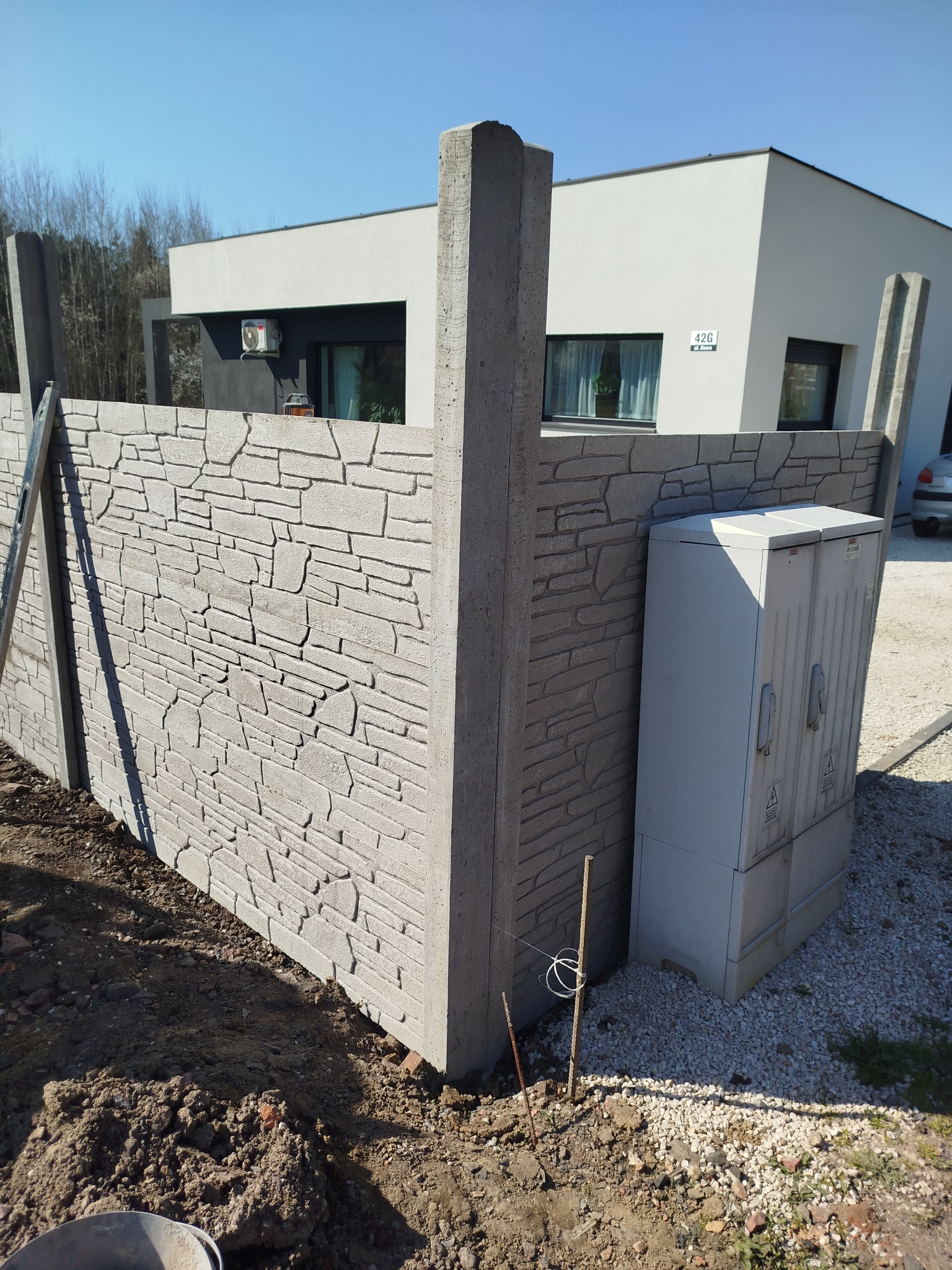 Montaż ogrodzenia betonowego z elementami imitującymi kamień, widoczna skrzynka elektryczna przy ogrodzeniu oraz nowoczesny dom w tle.