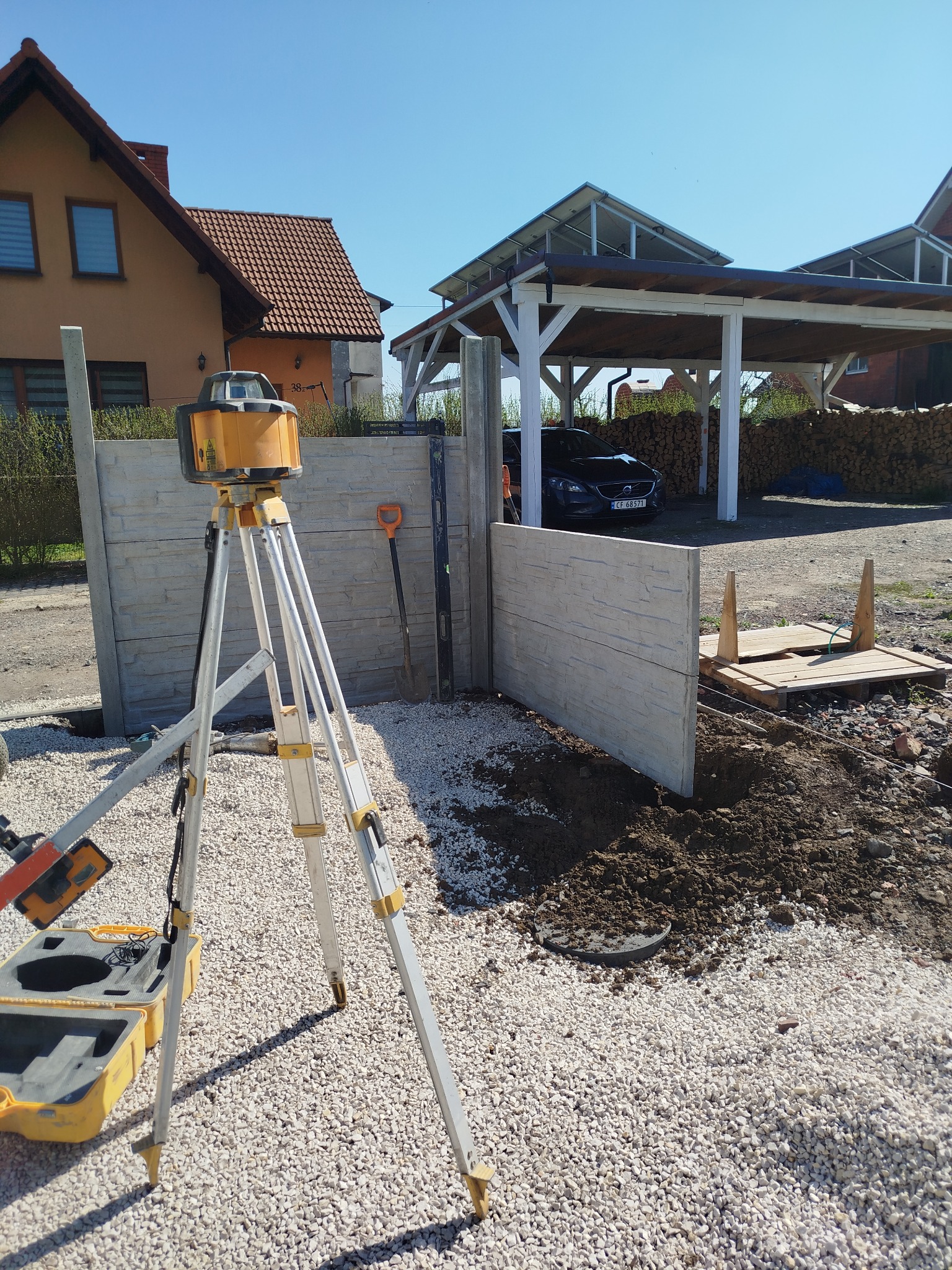 Montaż prefabrykowanych paneli ogrodzeniowych z użyciem niwelatora laserowego na trójnogu, widoczny wykop, podłoże wysypane drobnym kamieniem, w tle dom jednorodzinny i wiata na drewno.