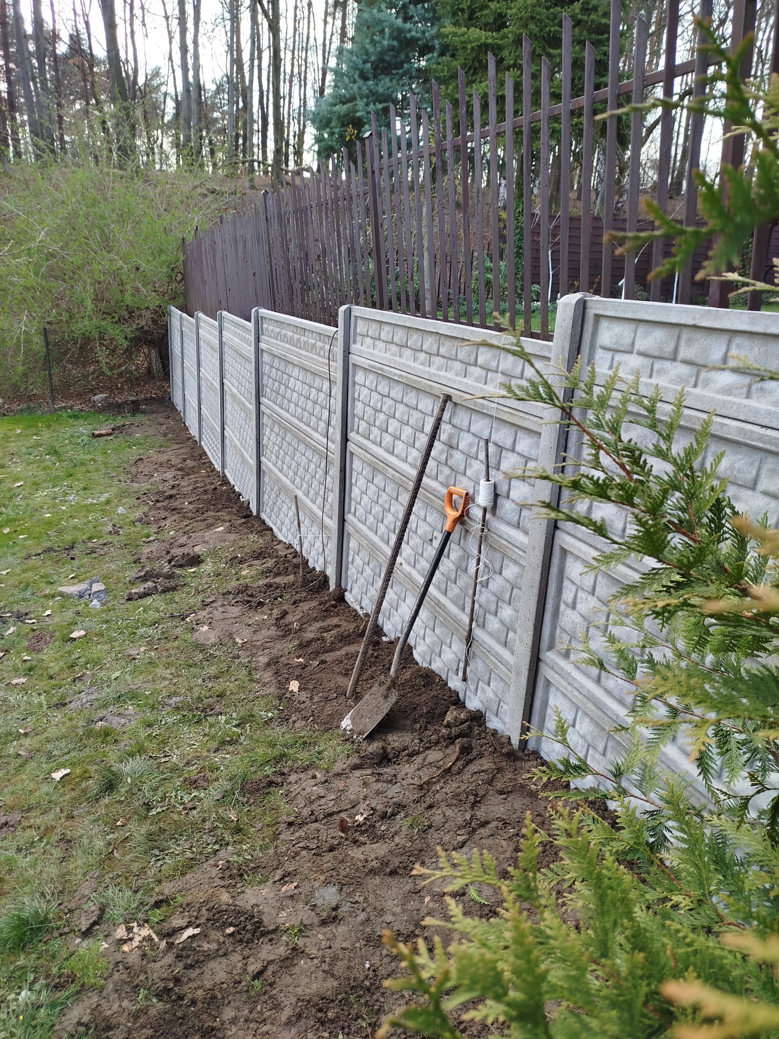 Szary, prefabrykowany mur z cegieł, w tle brązowe ogrodzenie z metalowych prętów, dwie łopaty oparte o przęsło, ziemia po montażu, trawa i krzewy.