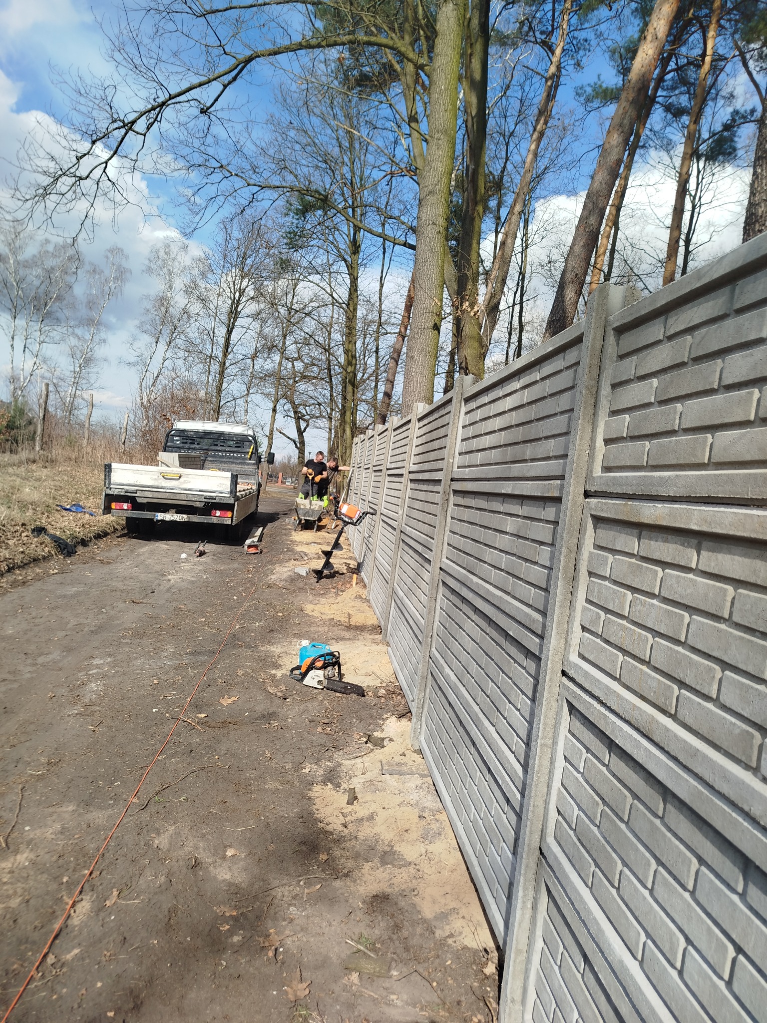 Montaż prefabrykowanego ogrodzenia betonowego z użyciem wiertnicy spalinowej, widoczny samochód dostawczy i dwóch pracowników w odzieży roboczej.