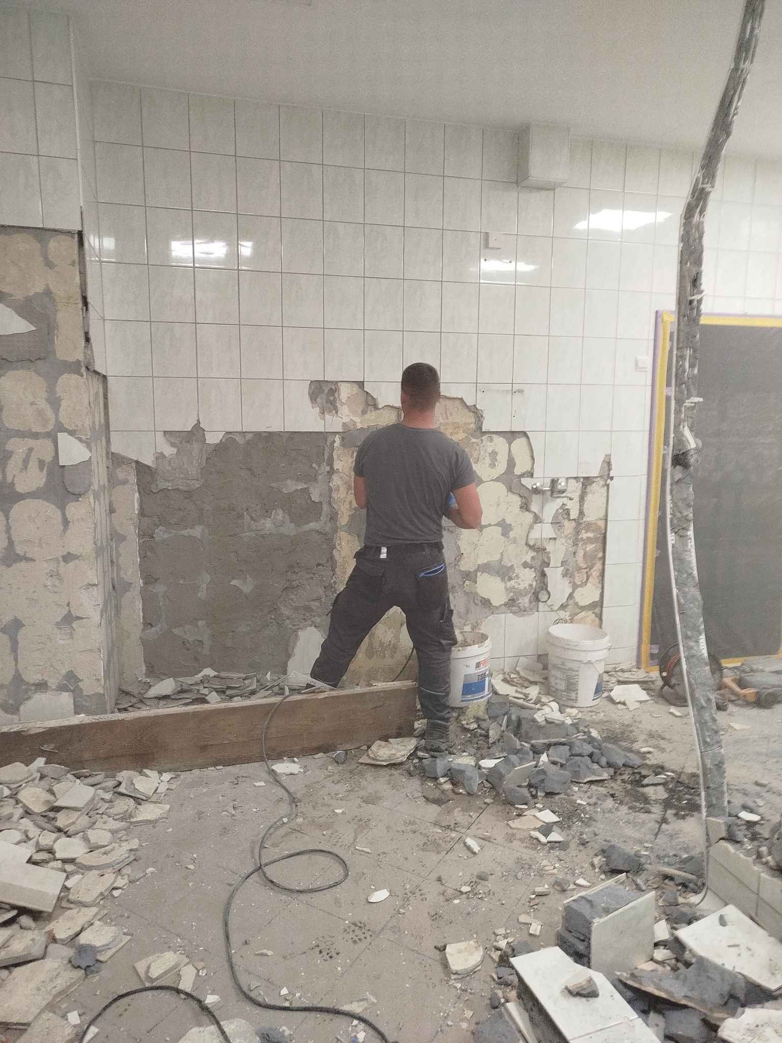 Demontaż płytek ściennych w łazience podczas remontu. Widoczny fragment ściany z resztkami kleju, gruz na podłodze i pracownik w trakcie pracy.