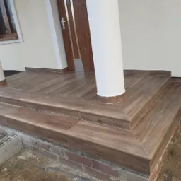 Wyłożone płytkami imitującymi drewno schody wejściowe z dwoma stopniami, z kolumną i fragmentem elewacji domu w tle.