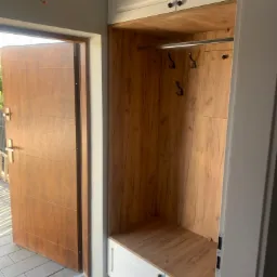 Wnęka wejściowa z drewnianym wykończeniem, wyposażona w wieszaki i szafki, z widocznymi drzwiami wejściowymi na zewnątrz.