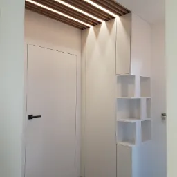 Biała szafa wnękowa z otwartymi półkami, wbudowana w przedpokoju, z drewnianym sufitem podwieszanym z oświetleniem LED, minimalistyczny design.