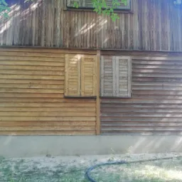 Fragment drewnianej elewacji budynku z dwoma oknami z okiennicami, gdzie jedna część ściany jest odnowiona i pomalowana, a druga w stanie surowym, prezentująca różnicę przed i po renowacji.