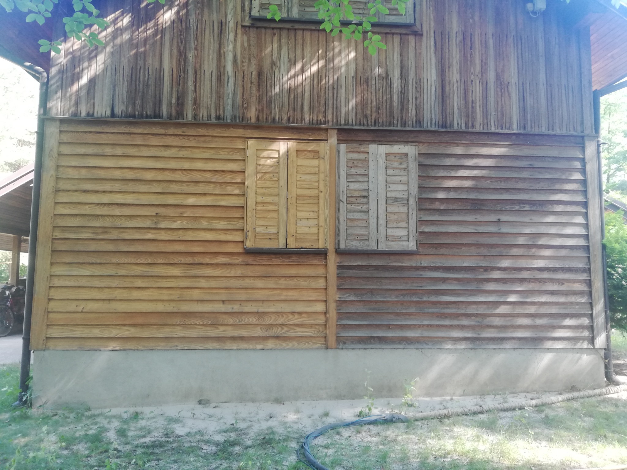 Fragment drewnianej elewacji budynku z dwoma oknami z okiennicami, gdzie jedna część ściany jest odnowiona i pomalowana, a druga w stanie surowym, prezentująca różnicę przed i po renowacji.