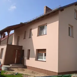 Dwukondygnacyjny dom z jasną elewacją i brązowymi akcentami, widoczny samochód osobowy i furgonetka w pobliżu, teren wokół domu w trakcie prac wykończeniowych.