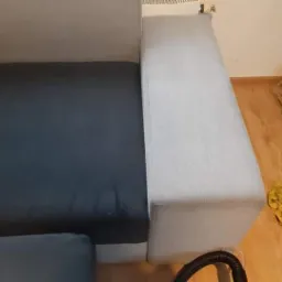 Fragment szarej sofy z ciemnym siedziskiem, widoczny wąż od odkurzacza i żółta ścierka na podłodze po prawej stronie.