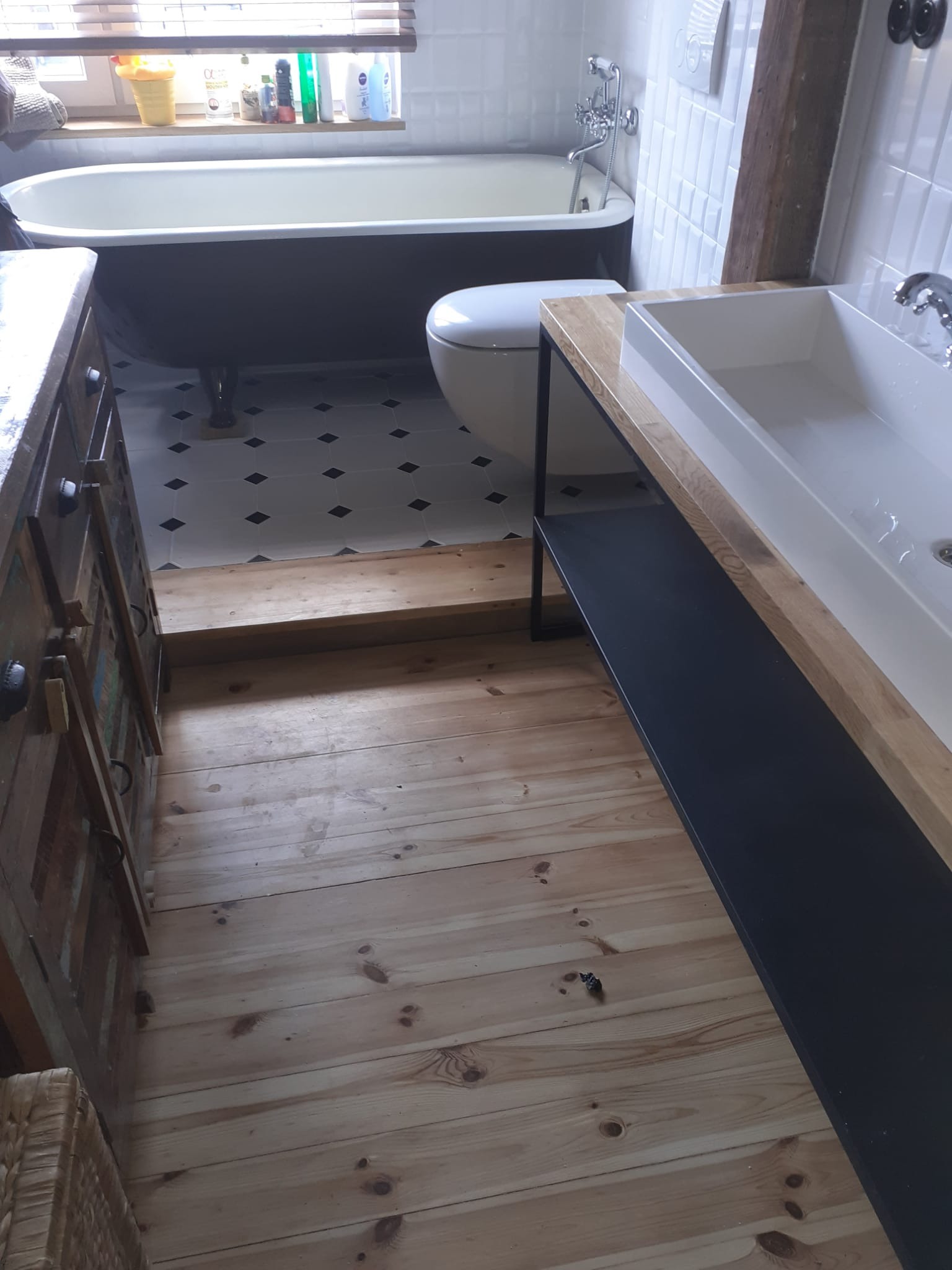 Nowoczesna łazienka z wolnostojącą wanną na nóżkach, toaletą, umywalką nablatową na drewnianym blacie wspartym na metalowym stelażu oraz drewnianą podłogą z desek.