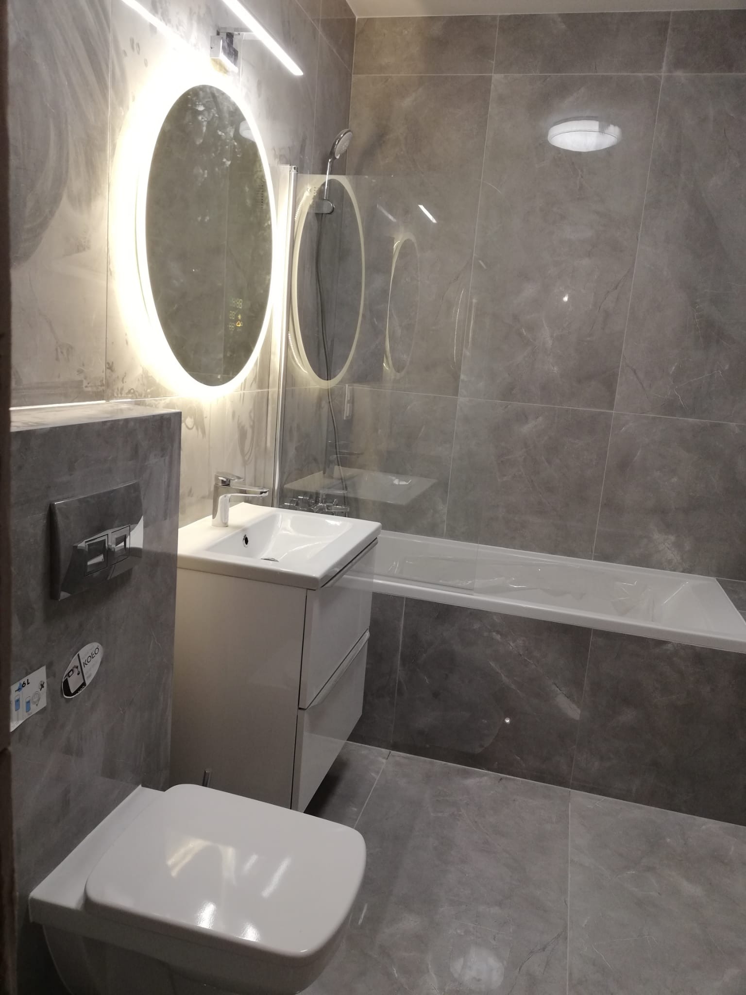 Nowoczesna łazienka z szarymi marmurkowymi płytkami, podświetlanym okrągłym lustrem, białą umywalką z szafką, podwieszaną toaletą i wanną za szklanym panelem.