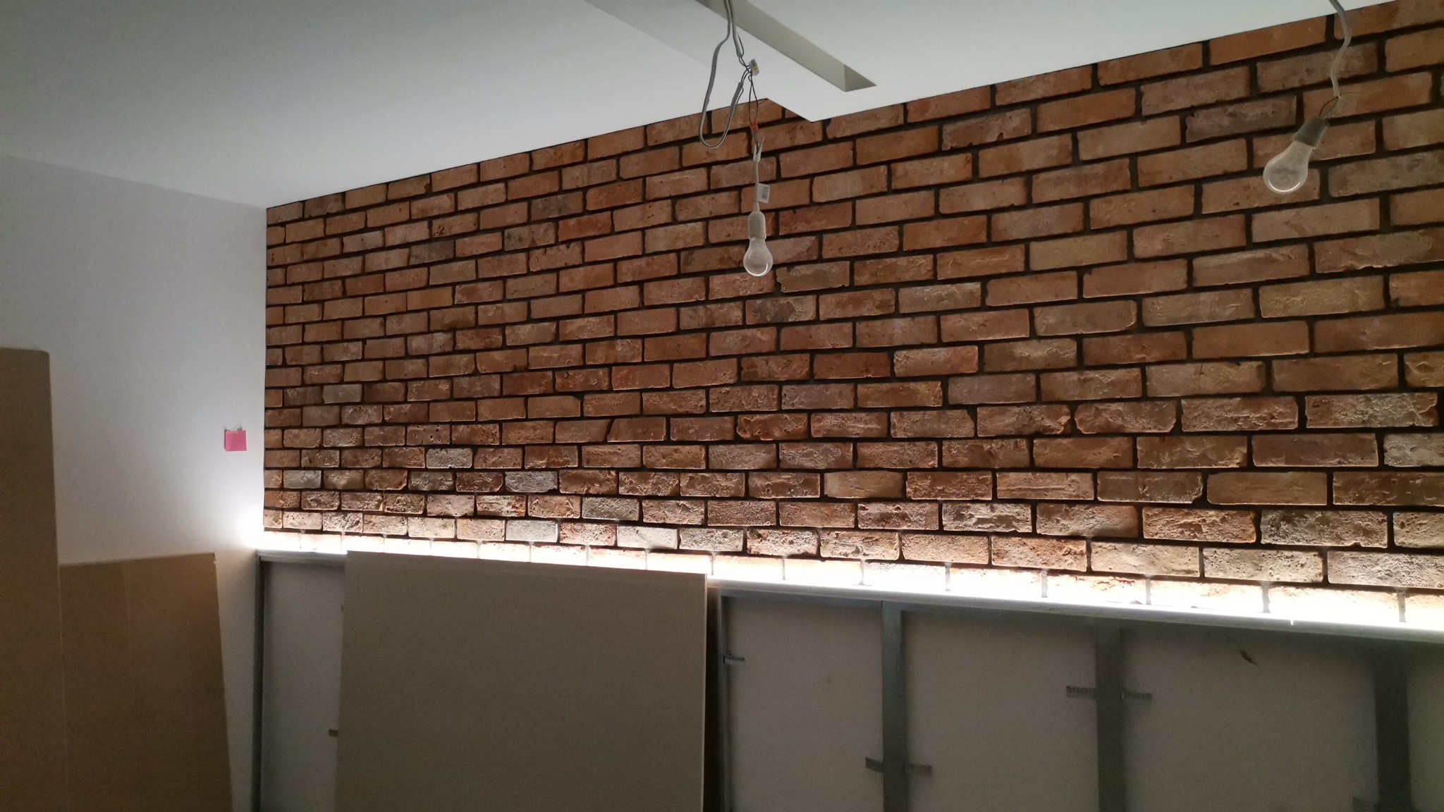 Montaż ściany z cegły dekoracyjnej z podświetleniem LED na stelażu z płyt kartonowo-gipsowych, widoczne wiszące żarówki i kartony.