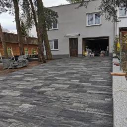 Ułożona kostka brukowa w szaro-czarnych odcieniach prowadząca do domu z garażem, widoczne drzewa i zaparkowany samochód w garażu.