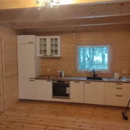 Wnętrze drewnianego domu z nową, białą zabudową kuchenną, widok na zewnątrz przez okno, podłoga i ściany z jasnego drewna, widoczny mop oparty o ścianę.