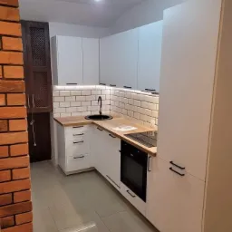 Nowoczesna kuchnia z białymi szafkami, drewnianym blatem i czarnymi detalami, oświetlona listwami LED pod szafkami wiszącymi, z fragmentem ceglanej ściany po lewej stronie.