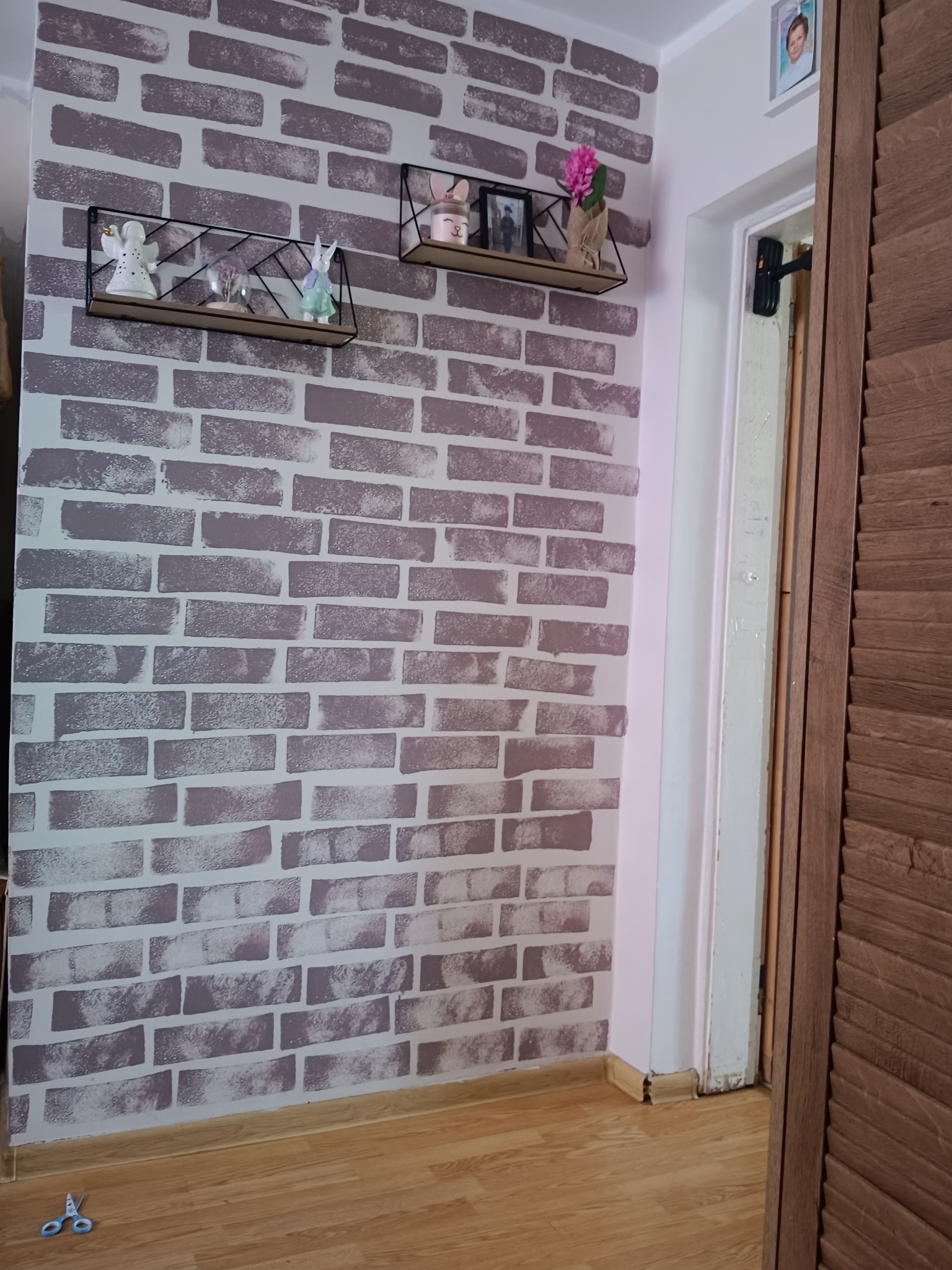 Ściana z tapetą imitującą cegłę, ozdobiona półkami z dekoracjami. Widoczne drzwi i podłoga. Na podłodze leżą nożyczki.