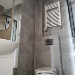 Nowoczesna łazienka z szarymi płytkami imitującymi marmur, białą umywalką z szafką, toaletą i grzejnikiem drabinkowym.