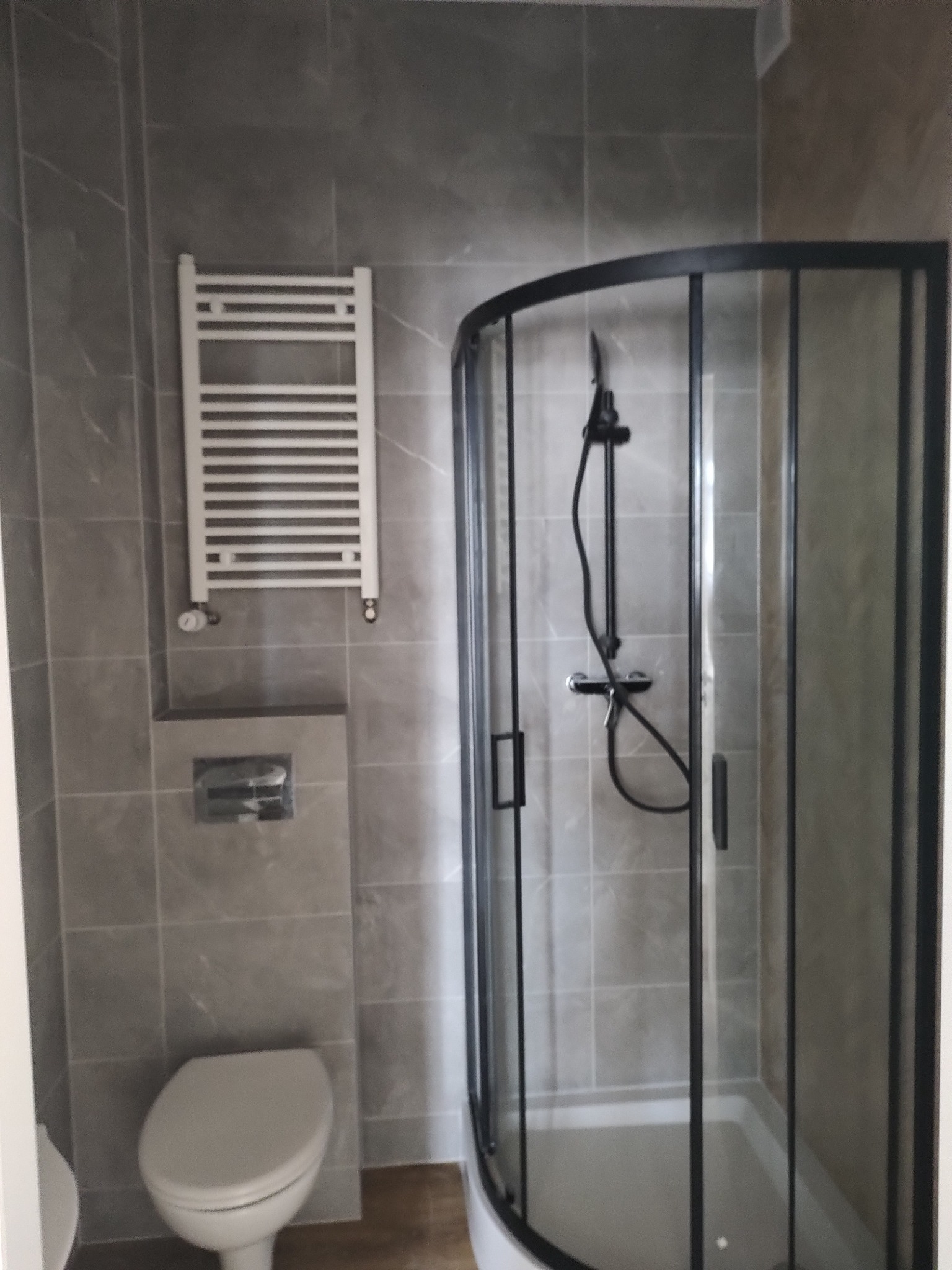 Wykończona łazienka z szarymi płytkami, kabiną prysznicową z czarną ramą, wiszącym grzejnikiem drabinkowym i podwieszaną toaletą.