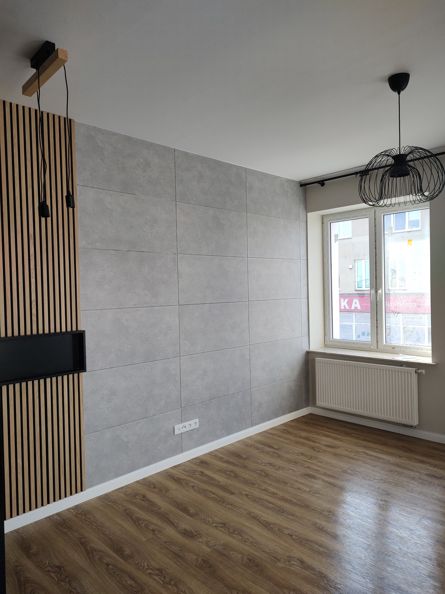 Pokój po remoncie: ściana wyłożona szarymi płytami, drewniane panele na podłodze, dekoracyjna ścianka z drewnianych listew z czarną wnęką, okno z grzejnikiem, nowoczesna lampa sufitowa.