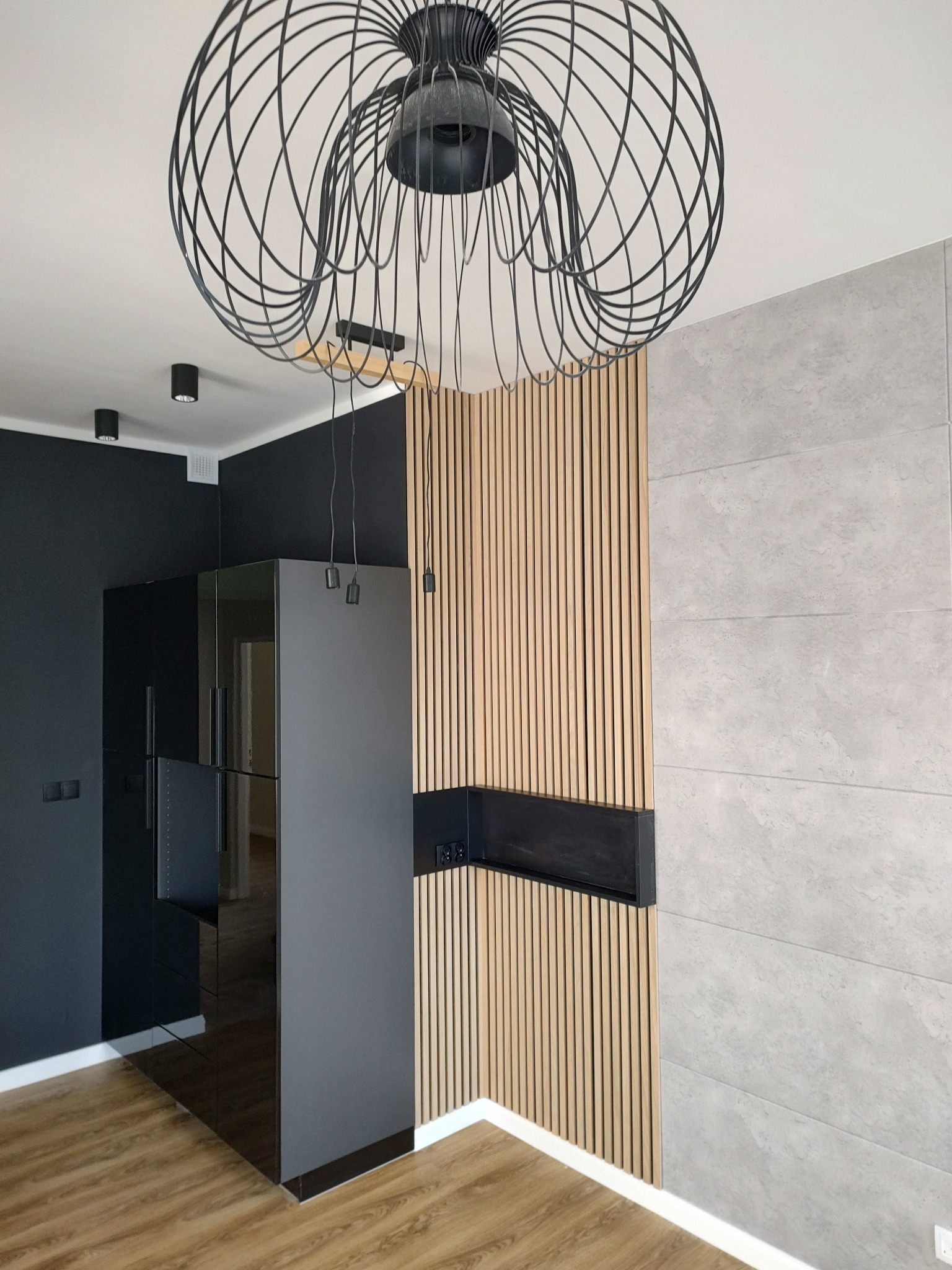 Nowoczesne wnętrze z drewnianymi lamelami na ścianie, szarą płytą imitującą beton, czarnymi szafkami i designerską lampą sufitową z metalowych prętów.