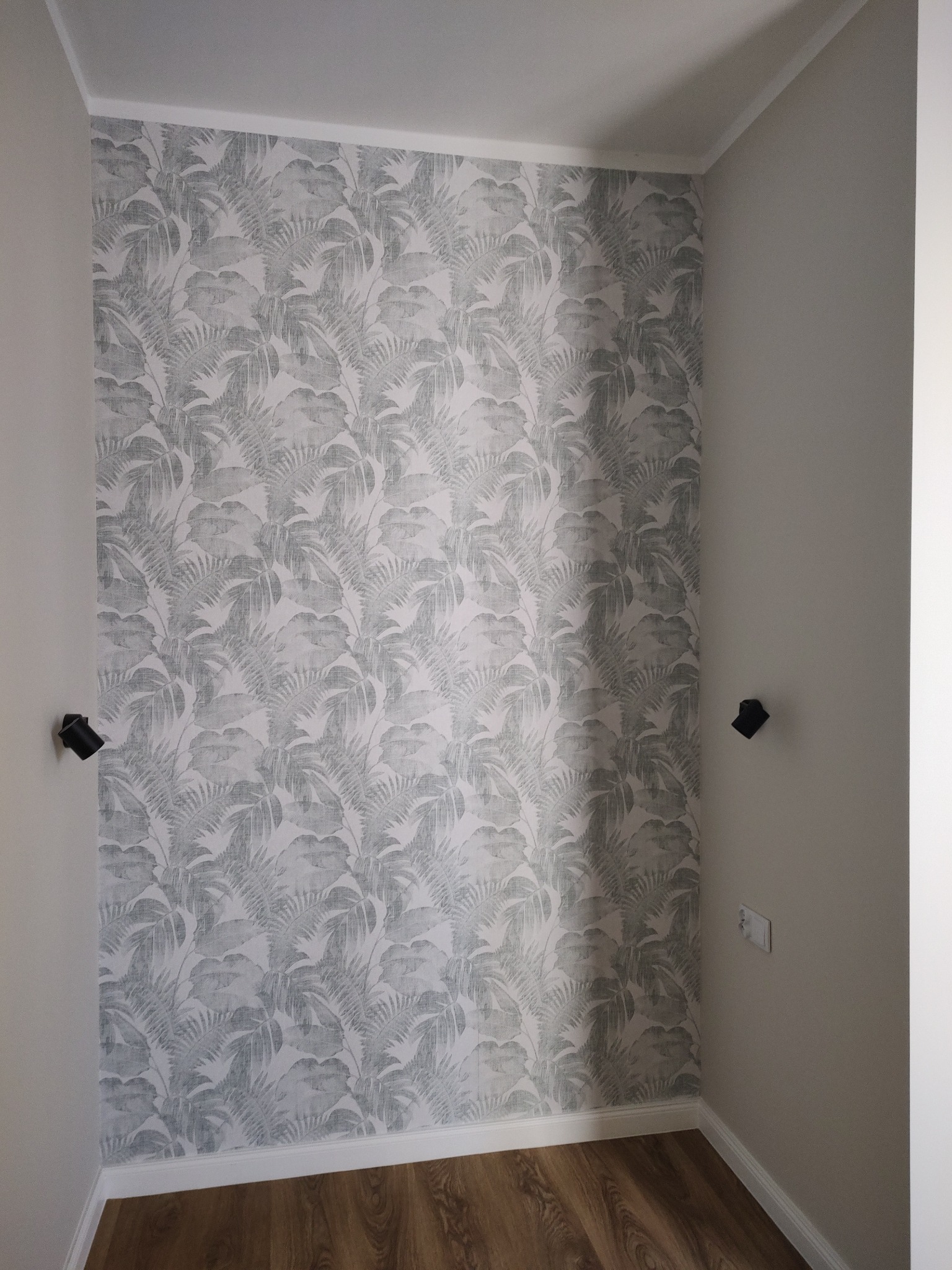 Pokój po odświeżeniu z tapetą w tropikalne liście i nową podłogą z paneli, widoczne czarne lampki ścienne i gniazdko elektryczne.