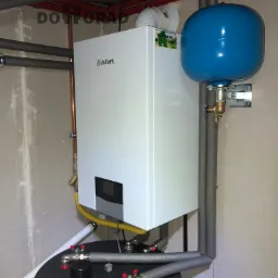 Biały wiszący kocioł gazowy Vaillant ecoTEC plus z niebieskim zasobnikiem 120 litrów, zainstalowany na tle surowej, betonowej ściany w piwnicy, widoczne rury miedziane i izolowane szare rury...