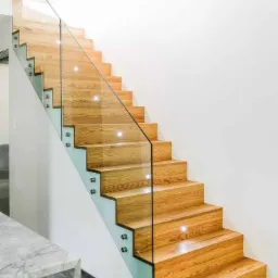 Drewniane schody z przezroczystą szklaną balustradą, mocowaną za pomocą metalowych uchwytów, w nowoczesnym wnętrzu z białą ścianą i marmurowym blatem w tle.