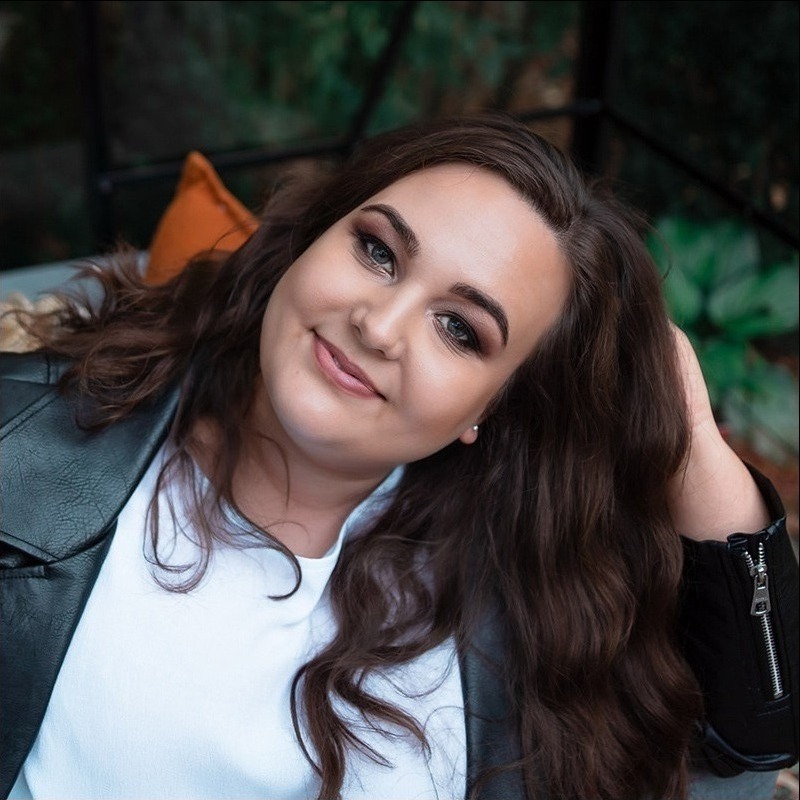 Portret uśmiechniętej brunetki w skórzanej kurtce, z delikatnym makijażem oczu i subtelnymi kolczykami, pozującej na tle zieleni.