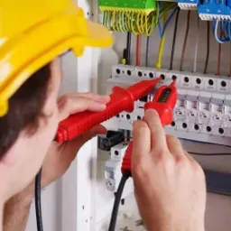 Elektryk w kasku, z czerwonym miernikiem, sprawdza instalację w rozdzielnicy. Widoczne przewody i bezpieczniki.