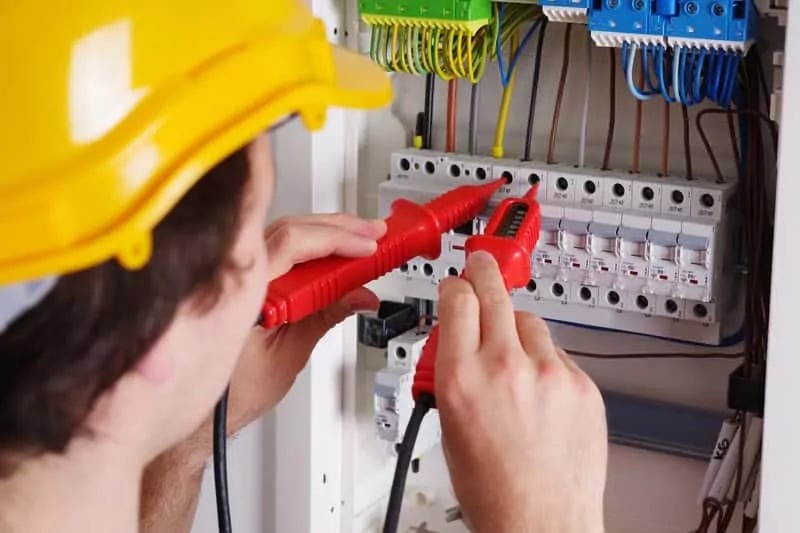 Elektryk w kasku, z czerwonym miernikiem, sprawdza instalację w rozdzielnicy. Widoczne przewody i bezpieczniki.
