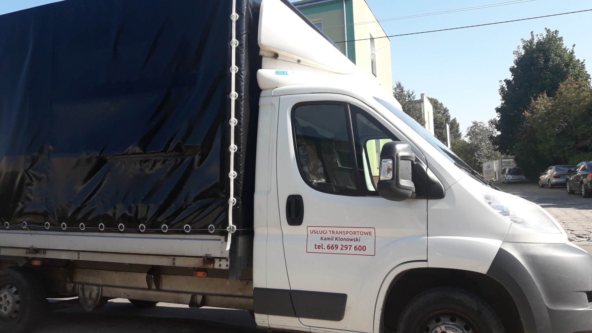 Biały samochód dostawczy z czarną plandeką i naklejką z danymi kontaktowymi firmy transportowej 'Usługi Transportowe Kamil Kłonowski' na drzwiach kabiny.