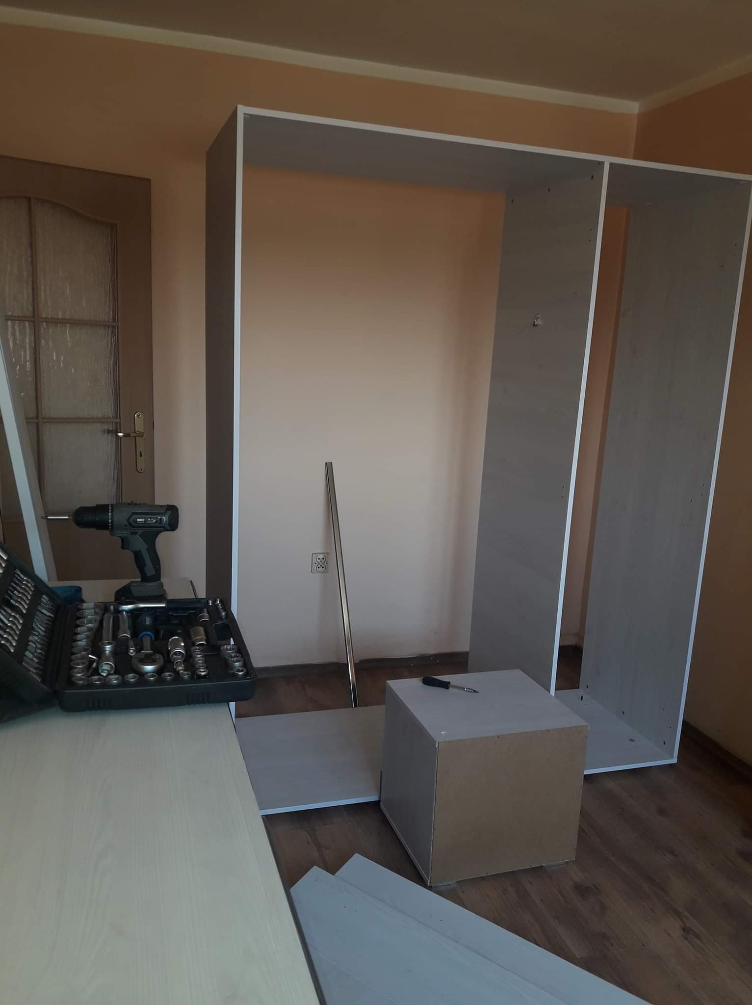 Montaż szafy: widoczne elementy konstrukcyjne, skrzynka z narzędziami, wkrętarka akumulatorowa i poziomica w pomieszczeniu o beżowych ścianach.