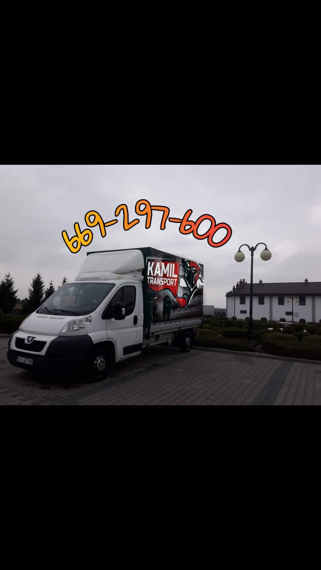 Biały bus dostawczy z nadrukiem 'Kamil Transport' i numerem telefonu, zaparkowany na brukowanym placu przed budynkiem z latarniami.