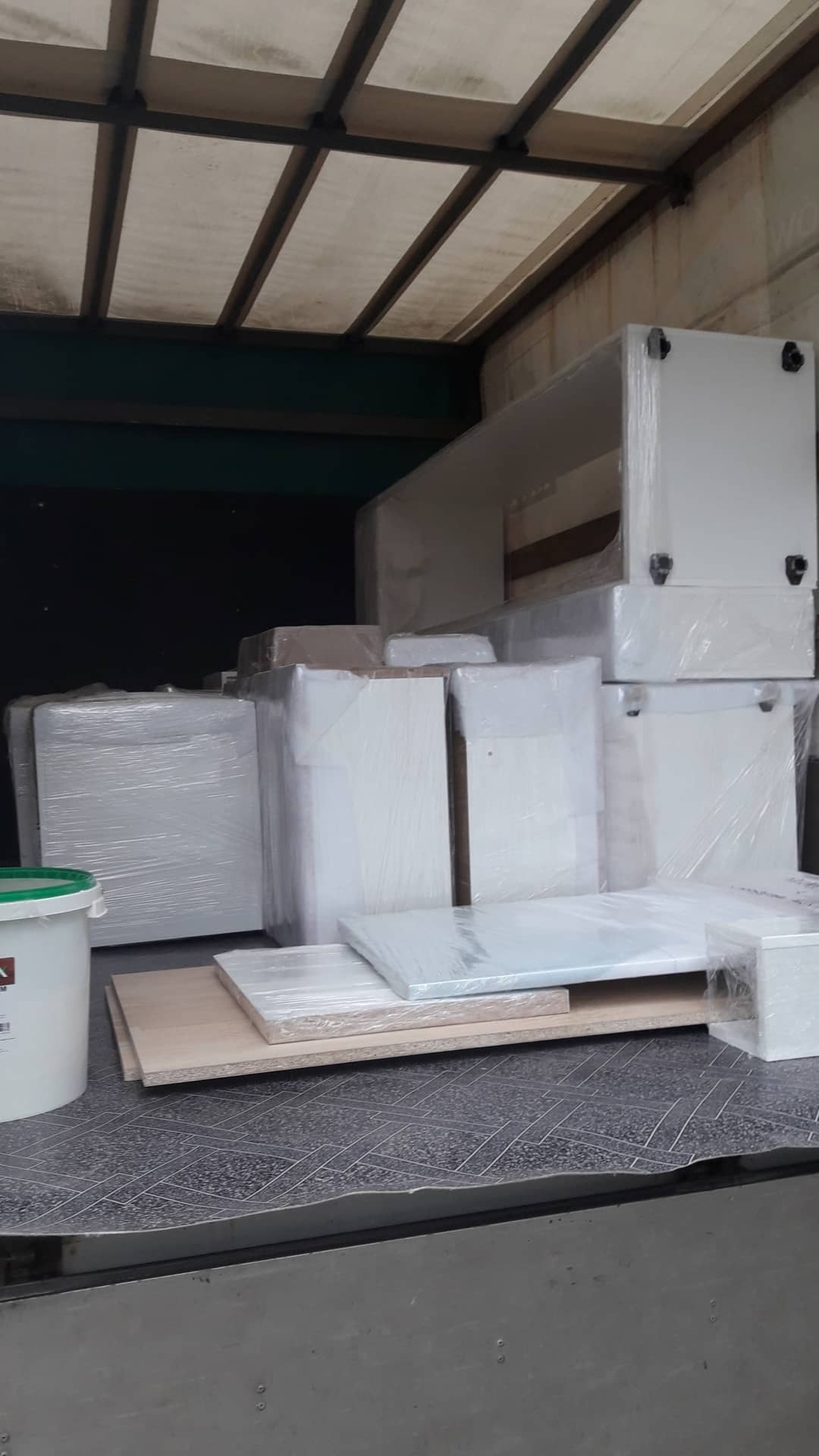 Meble zapakowane w folię stretch, częściowo ułożone na palecie, wewnątrz przestrzeni ładunkowej pojazdu dostawczego z widocznym zielonym wiadrem farby.