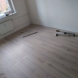 Usługi remontowo-wykończeniowe - Fragment układanej podłogi z paneli w jasnym odcieniu drewna, widoczny fragment listwy przypodłogowej i narzędzie do cięcia paneli.