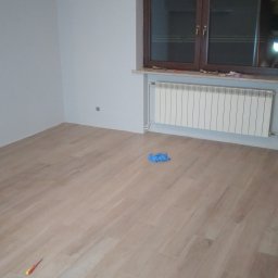 Usługi remontowo-wykończeniowe - Pokój po położeniu paneli podłogowych, widoczny grzejnik pod oknem, narzędzia pracy i niebieskie rękawice na podłodze.